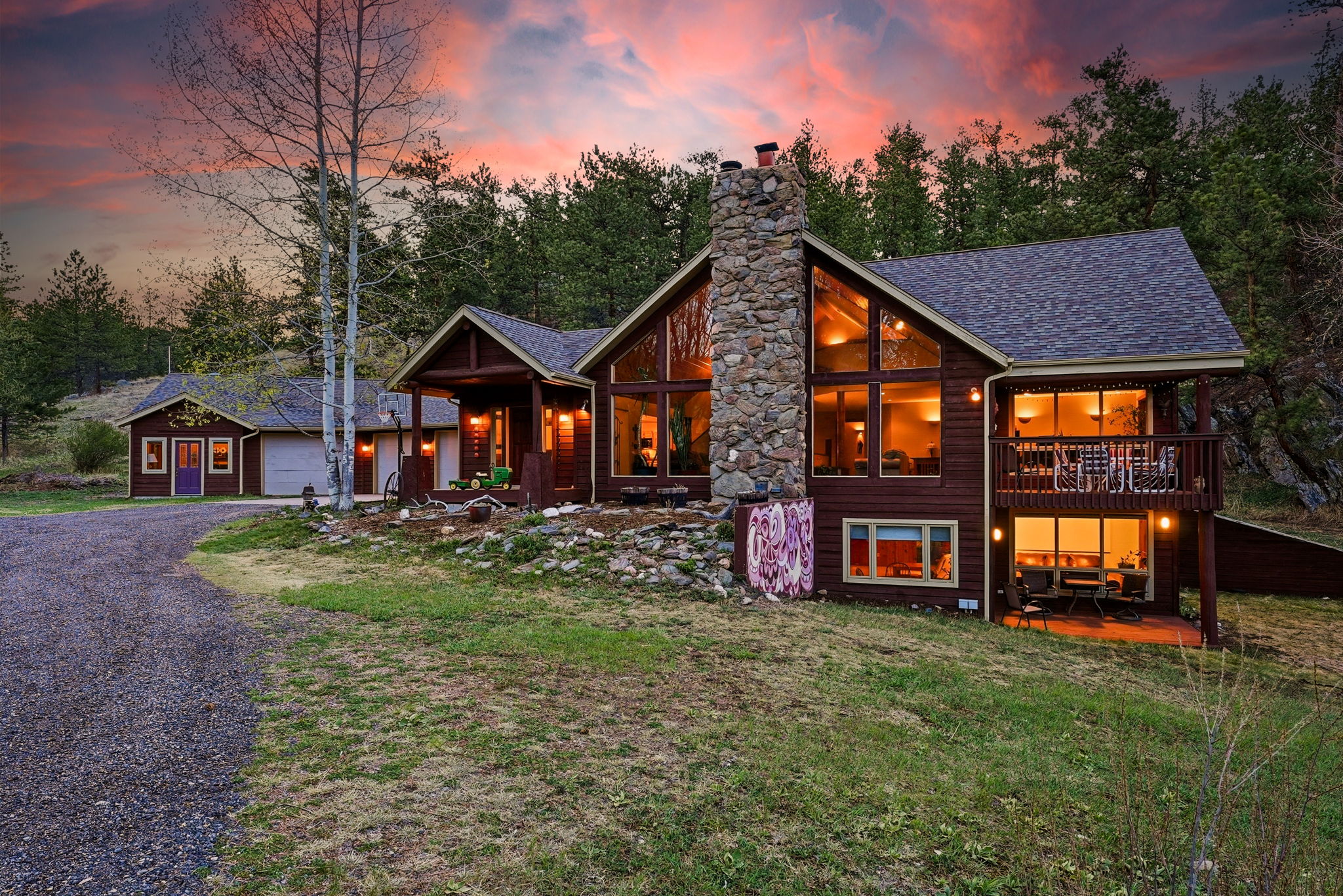 Rustic mountain-style exterior at Loveland acreage home for sale with stone fireplace, wooded surroundings, and warm evening lighting